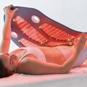 Woman lying on a bed holding a curved red LED light therapy panel against her abdomen. - SkinScience